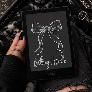 May include: A black e-reader with a white line-art graphic of a bow on the screen. Below the bow, the text reads "Brittany's Kindle" in a cursive font. The device has a black frame and the word "kindle" is printed in small letters at the bottom.