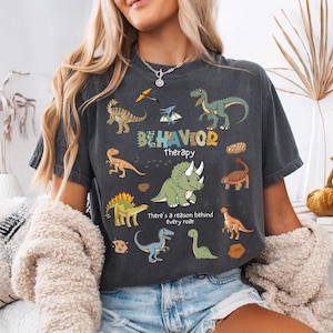 Op de afbeelding: Donkergrijs t-shirt met kleurrijke cartoon dinosaurussen en de woorden "BEHAVIOR therapy" en "there's a reason behind every roar". Het shirt heeft een vintage, gewassen uitstraling.