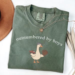 May include: Olive green t-shirt with the phrase "outnumbered by boys" in white. The shirt features a cartoon goose wearing a baseball cap and holding a baseball glove and a red truck. A gold necklace is draped across the shirt.