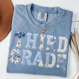 Könnte beinhalten: Ein hellblaues Comfort Colors T-Shirt mit den Worten "THIRD GRADE", die in verschiedenen blauen Blumen- und Mustermaterialien appliziert sind. Eine goldene Halskette mit Anhänger ist über dem Shirt drapiert.