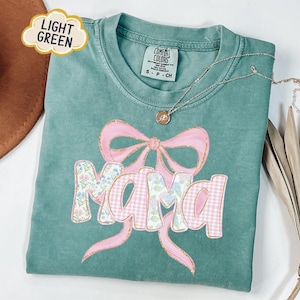 May include: Light green Comfort Colors t-shirt with a pink bow and the word "Mama" in a floral and gingham pattern. A gold necklace with a pendant is on the shirt. The shirt is folded and has a "Light Green" label.