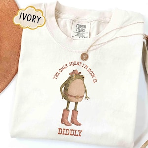 May include: Ivory-colored t-shirt with a cartoon frog wearing a cowboy hat and boots. The text reads "THE ONLY SQUAT I'M DOIN' IS DIDDLY" in brown. A gold necklace is draped on the shirt.