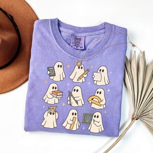 May include: A purple short-sleeved t-shirt featuring a graphic print of nine white ghosts engaged in various academic activities, including holding a calculator, pencil, geometry tools, books, a protractor, a pi symbol, a ruler, and a small chalkboard.