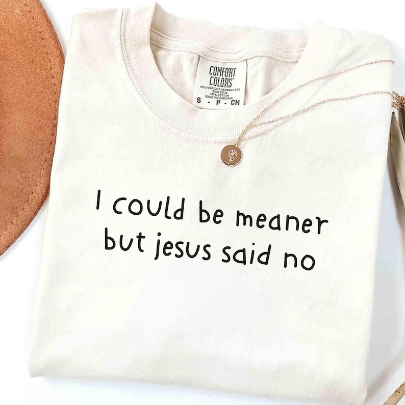 Pu&ograve; includere: T-shirt color crema con la scritta nera "I could be meaner but Jesus said no". Una collana dorata con un ciondolo &egrave; appoggiata sulla maglietta. La maglietta ha un'etichetta Comfort Colors.