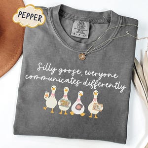 Puede incluir: Camiseta gris Comfort Colors con la frase "Silly goose, everyone communicates differently" en escritura blanca. La camiseta presenta un gráfico de cuatro gansos de dibujos animados, cada uno con diferentes herramientas de comunicación. También se ve un collar plateado con un colgante.