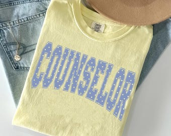 May include: A pale yellow short-sleeve t-shirt featuring the word COUNSELOR in a blue collegiate-style font with white polka dots. The shirt has a crew neckline and a Comfort Colors brand label on the inside collar area near the neck seam.