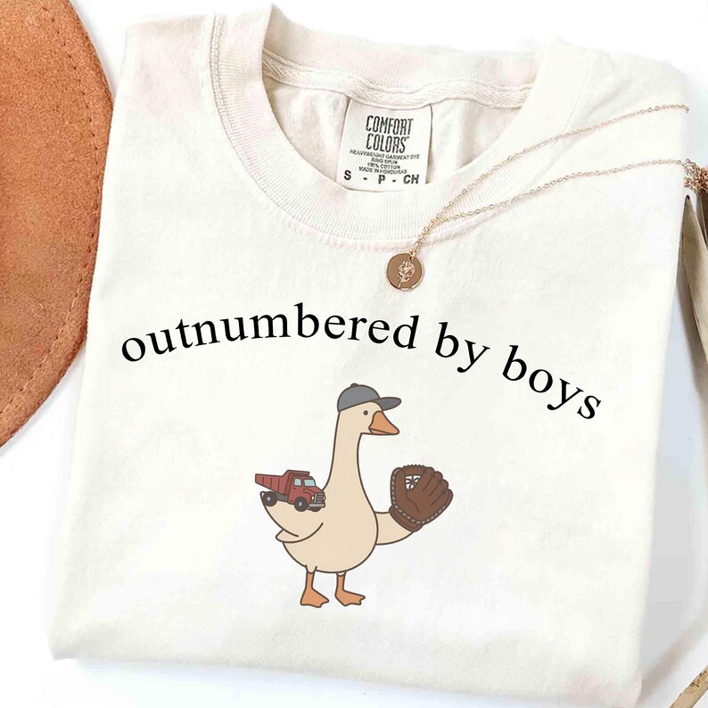 May include: Cream-colored t-shirt with the phrase "outnumbered by boys" in black. A cartoon goose wearing a baseball cap and glove is featured, along with a red truck graphic. A gold necklace is draped across the shirt.