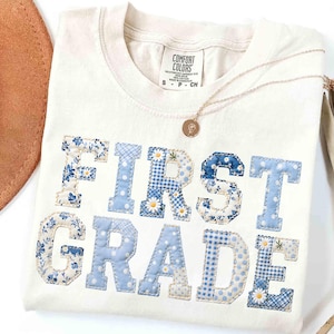 May include: Cream-colored t-shirt with the words "FIRST GRADE" appliquéd in blue floral, gingham, and polka dot patterns. A gold necklace with a pendant rests on the shirt. The shirt is from Comfort Colors.