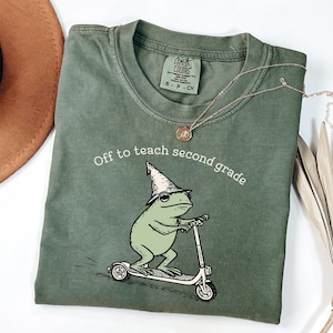 May include: Sage green t-shirt with the text "Off to teach second grade." A cartoon frog wearing a wizard hat rides a scooter. A gold necklace is draped across the shirt.