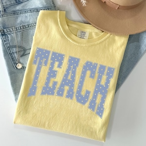 Könnte beinhalten: Ein hellgelbes Kurzarm-T-Shirt mit dem Wort TEACH in großen, hellblauen Blockbuchstaben mit weißem Punktmuster. Das Shirt hat einen Rundhalsausschnitt und ein Comfort Colors Markenetikett, das auf der Innenseite des Kragens aufgedruckt ist.