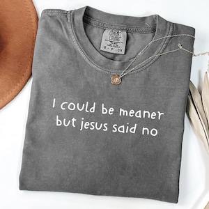 Pu&ograve; includere: Una maglietta grigia Comfort Colors con la scritta bianca "I could be meaner but Jesus said no". Una collana dorata con un ciondolo &egrave; sulla maglietta. Un cappello marrone &egrave; visibile in alto a sinistra.