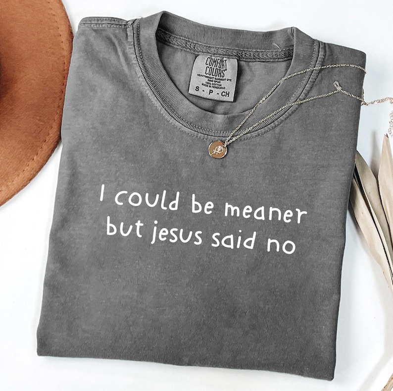 Pu&ograve; includere: Una maglietta grigia Comfort Colors con la scritta bianca "I could be meaner but Jesus said no". Una collana dorata con un ciondolo &egrave; sulla maglietta. Un cappello marrone &egrave; visibile in alto a sinistra.