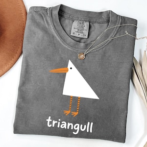Puede incluir: Una camiseta gris carbón de manga corta con un gráfico de pájaro geométrico blanco, pico naranja y patas naranjas a rayas. Debajo del pájaro, la palabra "triangull" está impresa en blanco. La camiseta tiene una etiqueta de Comfort Colors.