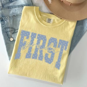 May include: A pale yellow short-sleeved t-shirt with a crew neckline and a Comfort Colors label. The shirt features large, light blue block letters with white polka dots spelling out the word FIRST, with the word grade in cursive script.