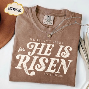 May include: Espresso-colored t-shirt with the text "HE IS NOT HERE for HE IS RISEN" in a distressed white font. The shirt includes the scripture reference Matthew 28:6. A silver necklace with a pendant is draped across the shirt.