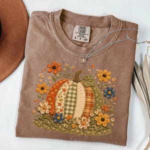 May include: A brown short-sleeve t-shirt featuring a central graphic of a pumpkin composed of various patterned fabric segments in orange, green, and cream. The pumpkin is surrounded by embroidered flowers in yellow, orange, and blue hues.