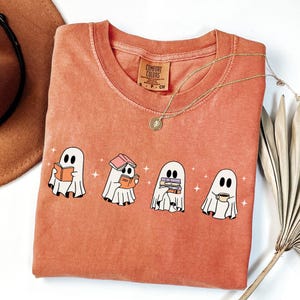 May include: A terracotta-coloured t-shirt featuring a graphic of four white ghosts. The first ghost reads an orange book, the second has a pink book on its head and an orange book in hand, the third holds a stack of books, and the fourth holds a mug.
