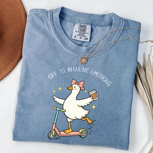 May include: A blue Comfort Colors t-shirt with the text "OFF TO REGULATE EMOTIONS". The graphic shows a cartoon duck with a pink bow riding a scooter and holding a coffee cup. A gold necklace is draped across the shirt.