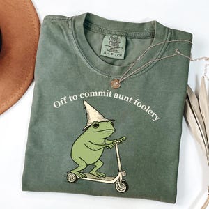 Op de afbeelding: Saliegroen t-shirt met de tekst "Off to commit aunt foolery" en een cartoon kikker met een tovenaars hoed op een scooter. Een zilveren ketting met een hanger ligt over het shirt.