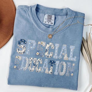 May include: A light blue Comfort Colors t-shirt with the words "SPECIAL EDUCATION" appliqued in a patchwork design of floral and gingham patterns. A gold necklace with a pendant rests on the shirt. A brown hat is visible in the background.
