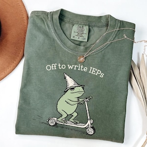 Könnte beinhalten: Olivgrünes T-Shirt mit dem Text "Off to write IEPs" und einem Cartoon-Frosch mit Zaubererhut, der einen Roller fährt. Eine goldene Halskette und ein brauner Hut sind ebenfalls zu sehen.