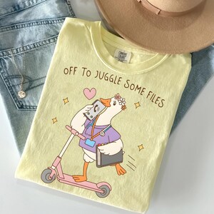May include: A pale yellow t-shirt featuring a graphic of a white goose wearing a purple shirt, a stethoscope, and a flower crown. The goose is riding a pink scooter while holding a phone and a folder. The text reads, "OFF TO JUGGLE SOME FILES."