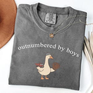 May include: Dark gray Comfort Colors t-shirt with the phrase "outnumbered by boys" in white. A cartoon goose wearing a baseball cap and holding a baseball glove and a red truck is printed on the front. A silver necklace with a pendant is also visible.
