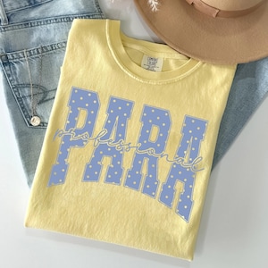 May include: A pale yellow short-sleeve t-shirt featuring the word PARA in large, light blue block letters with white polka dots, overlaid with the word professional in a cursive blue script font. The shirt has a crew neckline and a tag.