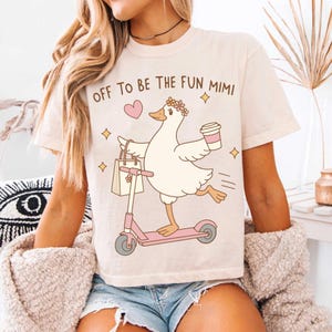 May include: A cream-coloured t-shirt with the text "OFF TO BE THE FUN MIMI." The design shows a cartoon duck on a pink scooter, carrying a shopping bag and a coffee cup. The duck is wearing a floral crown.
