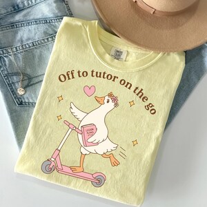 以下が含まれることがあります： 淡い黄色のTシャツ。花冠をかぶり、ピンクのキックボードに乗り、「LESSON PLANS」と書かれたピンクの本を持つ白いアヒルのグラフィックが描かれています。アヒルの上には茶色で「Off to tutor on the go」という文字がプリントされています。