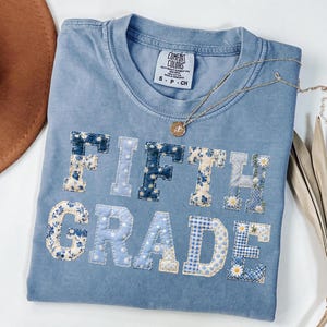 May include: A light blue Comfort Colors t-shirt with the words "FIFTH GRADE" appliqued on the front in various floral and patterned fabrics. A gold necklace with a pendant is draped across the shirt.