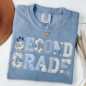 May include: A light blue Comfort Colors t-shirt with the words "SECOND GRADE" appliqued in various blue floral and gingham patterns. A gold necklace with a pendant rests on the shirt.