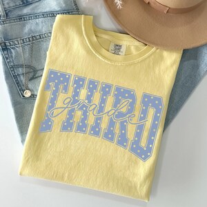 May include: A pale yellow short-sleeved t-shirt featuring the text "THIRD grade" in a light blue block font with white polka dots. The shirt has a Comfort Colors label at the inner neckline. The garment is displayed alongside blue denim fabric.