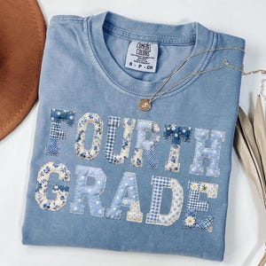Può includere: Una maglietta Comfort Colors azzurra con le parole "FOURTH GRADE" applicate in vari tessuti floreali e fantasia blu. Una collana d'oro con un ciondolo poggia sulla maglietta. Un cappello marrone è visibile nell'angolo in alto a sinistra.