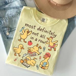 May include: A pale yellow t-shirt featuring a graphic of five cartoon ducks with school supplies and the text "most definitely not all in a row". The design includes a duck with a ruler, a duck with a pencil, a duck on books, a duck reading, and a duck wearing a "TEACH" cap.
