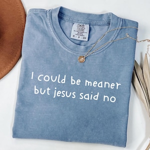 Può includere: Una maglietta blu Comfort Colors con la scritta bianca "I could be meaner but jesus said no". Una collana d'oro con un ciondolo è drappeggiata sulla maglietta. Un cappello marrone è visibile in alto a sinistra.