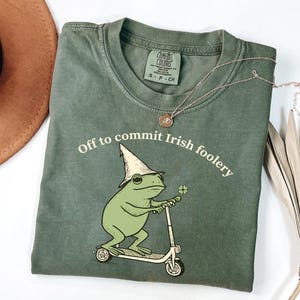 May include: Green t-shirt with the text "Off to commit Irish foolery" and a cartoon frog wearing a hat and riding a scooter. The frog holds a four-leaf clover. A silver necklace with a pendant is also visible.