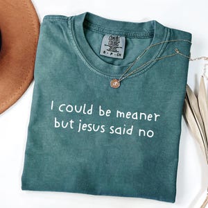 Pu&ograve; includere: Una maglietta Comfort Colors verde acqua con la scritta bianca "I could be meaner but jesus said no". Una collana d'oro con un ciondolo &egrave; drappeggiata sulla maglietta. La maglietta &egrave; piegata su una superficie bianca.