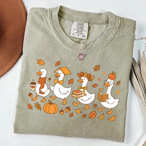 May include: A sage green t-shirt featuring a graphic of four white geese in autumn attire. The geese are depicted with a coffee cup, a book on their head, a leaf crown, and an apple on their head. The design includes orange pumpkins and leaves.