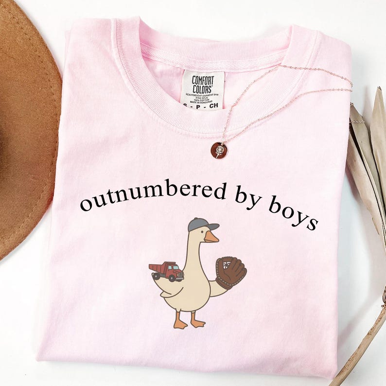 May include: A light pink t-shirt with the phrase "outnumbered by boys" printed in black. The shirt features a cartoon goose wearing a baseball cap and holding a baseball glove, with a red truck on its back. A necklace is also visible.