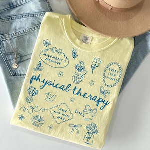 May include: A pale yellow t-shirt featuring blue line art illustrations of flowers, butterflies, a watering can, and a basket. The text reads "MOVEMENT IS MEDICINE," "physical therapy," "EVERY STEP COUNTS," and "GROW YOUR OWN WAY" in a blue script font.