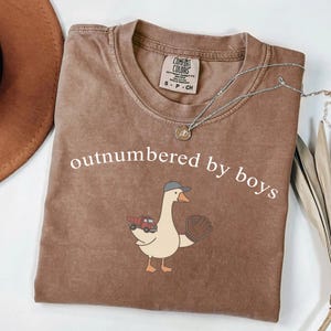 May include: Brown Comfort Colors t-shirt with the text "outnumbered by boys". The shirt features a cartoon goose wearing a baseball cap and holding a baseball glove and a red truck.