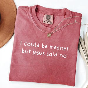 Pu&ograve; includere: T-shirt Comfort Colors rosa antico con la scritta bianca "I could be meaner but jesus said no". Una collana dorata con un piccolo ciondolo &egrave; drappeggiata sulla maglietta. Taglia S.