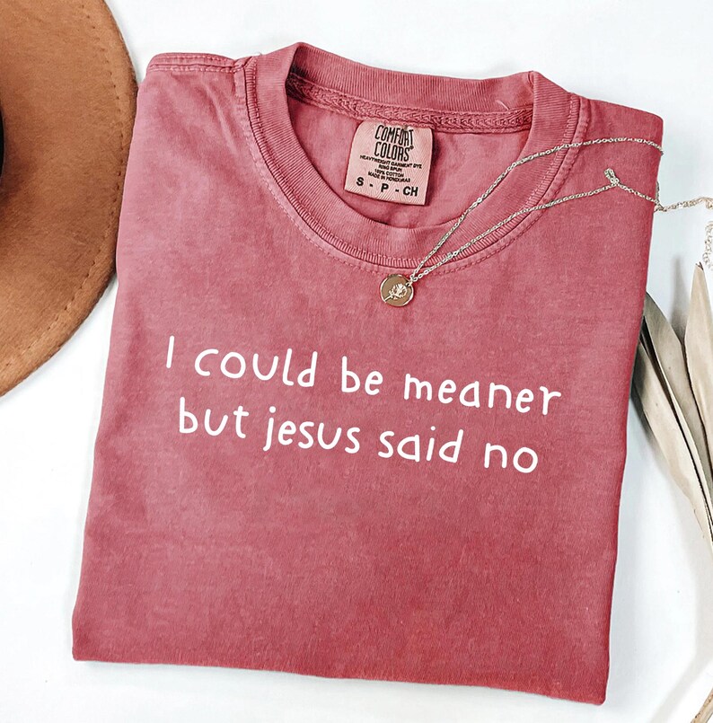 Pu&ograve; includere: T-shirt Comfort Colors rosa antico con la scritta bianca "I could be meaner but jesus said no". Una collana dorata con un piccolo ciondolo &egrave; drappeggiata sulla maglietta. Taglia S.