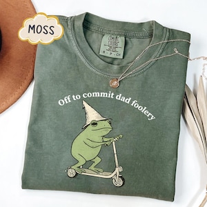 Op de afbeelding: Mosgroen t-shirt met de witte tekst "Off to commit dad foolery". Het ontwerp toont een cartoon kikker met een tovenaars hoed op een scooter. Een gouden ketting ligt over het t-shirt.