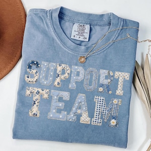 May include: A light blue t-shirt with the words "SUPPORT TEAM" appliqued on the front in various floral and patterned fabrics. A gold necklace with a small pendant rests on the shirt. The shirt has a Comfort Colors tag.