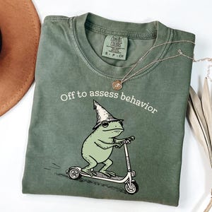 May include: Olive green t-shirt with the phrase "Off to assess behavior" above a cartoon frog riding a scooter. The frog wears a wizard hat. A gold necklace is draped across the shirt.