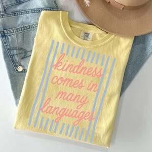 May include: A pale yellow short-sleeve t-shirt featuring a graphic design with vertical light blue stripes and the pink cursive text "kindness comes in many languages" centered on the front. The shirt has a crew neckline and a Comfort Colors label.