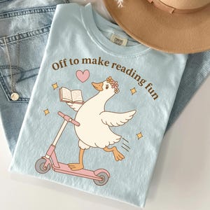 May include: A light blue t-shirt featuring a graphic of a white goose wearing a flower crown, riding a pink scooter, and holding an open book. The text "Off to make reading fun" is printed in brown above the goose, accompanied by a pink heart.