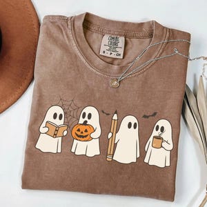 Op de afbeelding: Een bruin t-shirt met korte mouwen en een afbeelding van vier witte geesten. Eén geest houdt een open boek vast, één een pompoen, één een potlood en één een koffiemok. Boven de geesten zijn een spinnenweb en twee vleermuizen afgebeeld. Het t-shirt heeft een label van het merk Comfort Colors.
